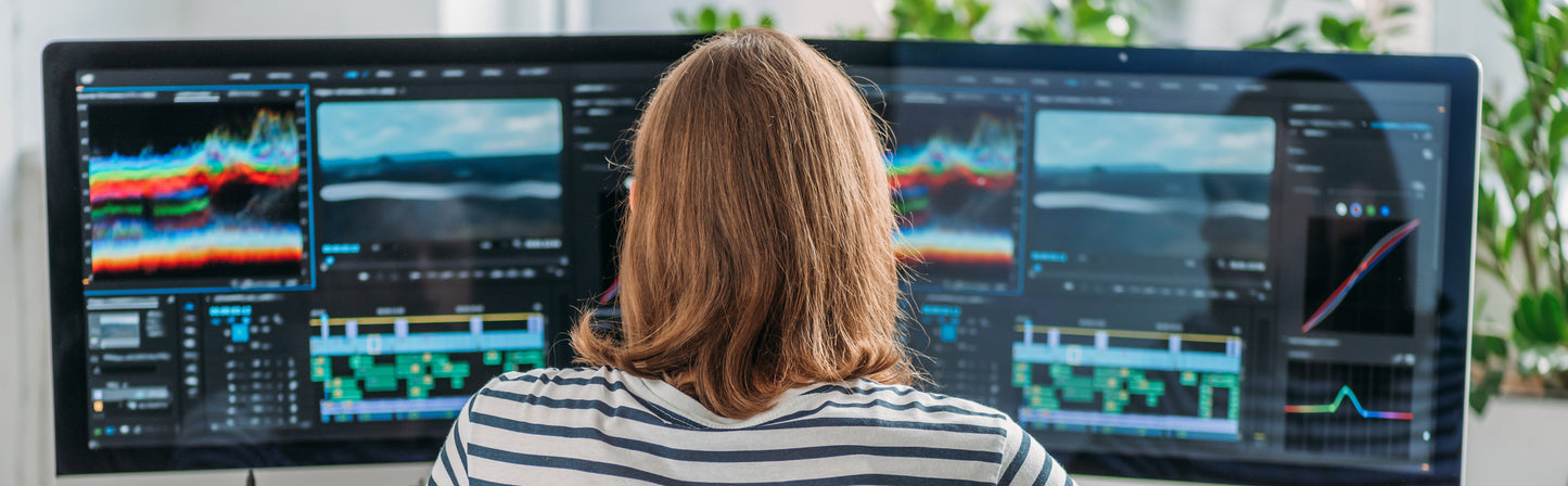 Person with shoulder-length hair sitting at a desk, editing video footage on a large dual-monitor setup with multiple editing panels, waveforms, and timelines displayed on the screen.